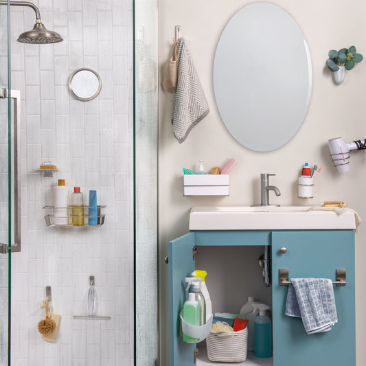 Ein Badezimmer mit einer Glasdusche auf der linken Seite, einem blauen Waschtisch, einem weißen Waschbecken, einem ovalen Spiegel und organisierten Toilettenartikeln. Handtücher hängen ordentlich am 3M Command™ Großer Badezimmer-Doppelhaken Nickel satiniert von 3M Deutschland GmbH nebenan.