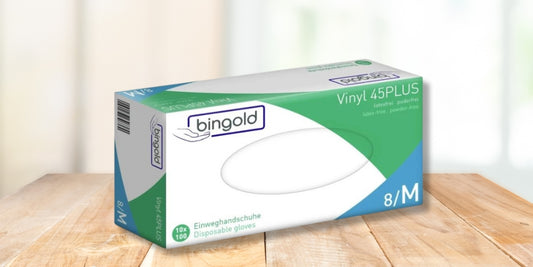 Eine Schachtel mit Bingold Vinyl 45PLUS Einweghandschuhen, Größe 8/M, steht auf einer Holzfläche. Die Schachtel ist mit grünen und blauen Akzenten versehen und enthält 10 Packungen mit je 100 Handschuhen.