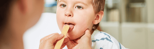 Kind mit geöffnetem Mund bei Untersuchung, während eine Person mit Holzspatel in den Rachen schaut, Nahaufnahme in Innenraum