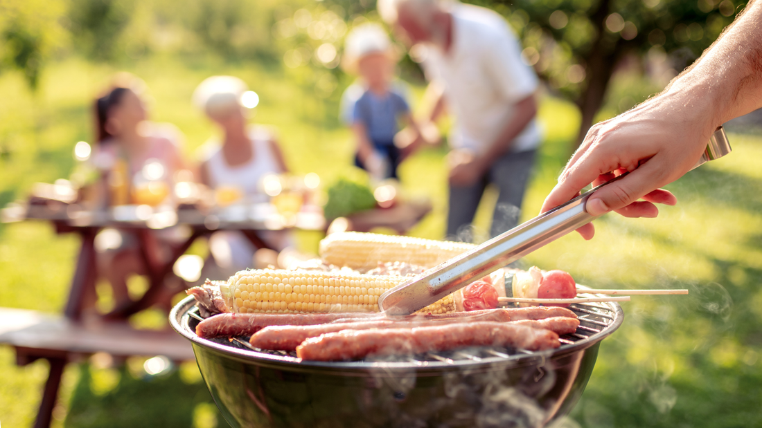 Familie die zusammen im Garten auf einer Bank sitzen und jemand im Vordergrund Würste und Mais auf einem Grill brät