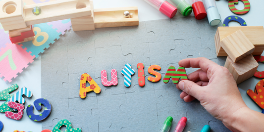 Bunte Buchstaben formen das Wort „Autism“ auf Puzzlefläche, Hand legt letzten Buchstaben, Spielzeug im Hintergrund
