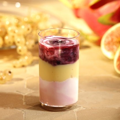 A Starpak 30 finger food cup, PS 60 ml, Ø 4 cm, 7.5 cm clear glass, is filled with a layered dessert in pink, yellow, and purple and garnished with purple sauce. In the background: light berries as well as fig and dragon fruit slices.