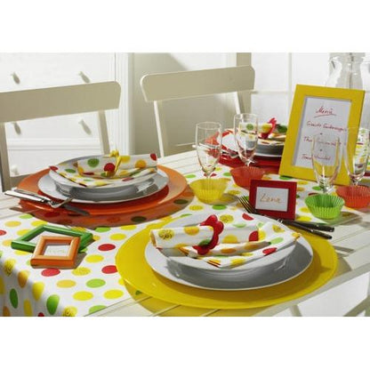 A table for two with yellow and orange placemats, white plates, PAPSTAR 20 napkins 33x33 cm from PAPSTAR GmbH, glasses, framed place cards, a red-yellow-green dotted tablecloth and decoration, and a framed menu in the background.