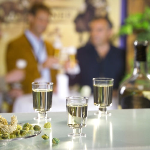 The Starpak 20 stem glasses for white wine made of crystal-clear polystyrene stand on a table with olives and a bottle, while three people chat over drinks in the background.