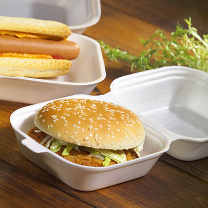 A hamburger in a sesame bun with lettuce and patty, served in a Starpak 100 hamburger box with a hinged lid, XPS, 7.5 x 12 x 12 cm white – ideal for catering, with hotdogs nearby and leafy greens on a wooden surface.