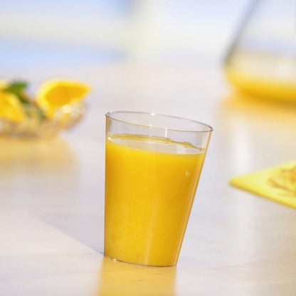A Starpak 10 drinking cup, PS 0.2l crystal clear, filled with orange juice stands on a light table. In the background, blurred orange slices and a jug can be seen.