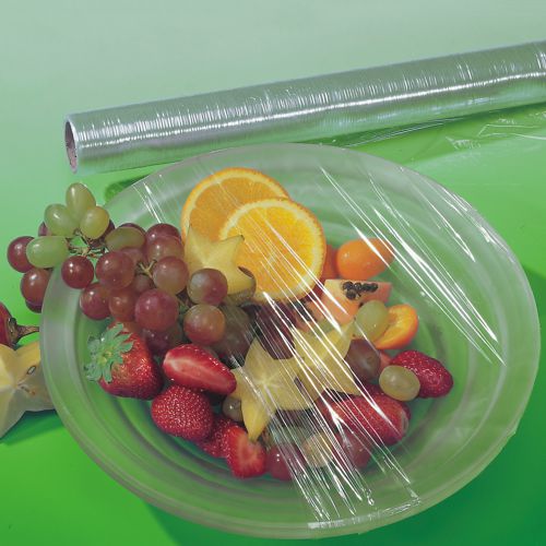 A transparent sheet of Starpak cling film (30 m x 29 cm) covers a plate with mixed fruit. In the background, on a green surface, the tear-resistant Starpak cling film roll in its folding box (1 piece) can be seen.