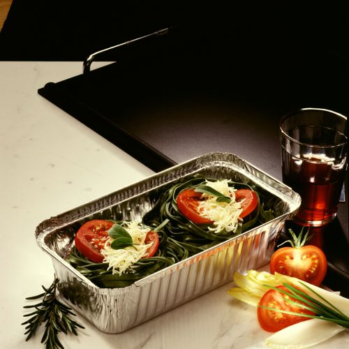 A Starpak 25 tray, aluminum + insert lid, PP-coated tray contains uncooked green pasta nests with cheese, tomato slices, and basil. Next to it: a glass of red drink, tomatoes, endive, and rosemary. Ideal for catering or take-away.