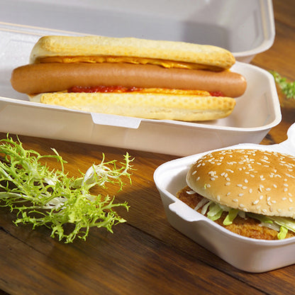 A hot dog with ketchup and mustard is served in a Starpak 100 all-purpose box with a hinged lid XPS 1800 ml, next to it a sesame hamburger with lettuce in another Starpak box. On the wooden table lies some curly lettuce leaf.