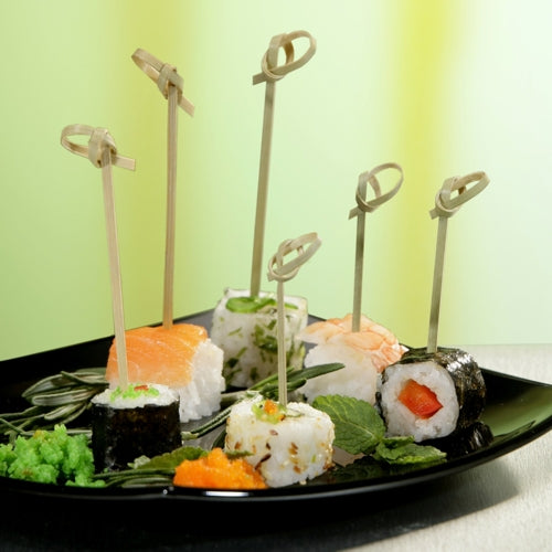 A black plate with various sushi, each piece skewered with PAPSTAR 40 finger food picks "pure" from PAPSTAR GmbH and garnished with herbs and fish roe, is elegantly presented against a background with a green gradient.