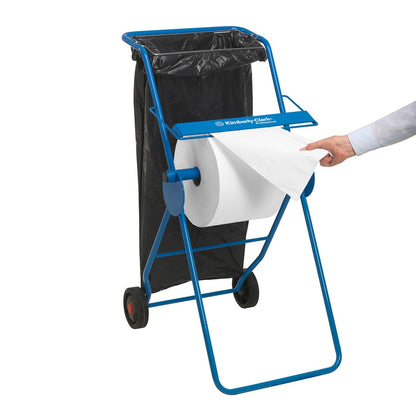 A blue metal dispenser holds a WypAll® L10 paper towel for surfaces - large roll (38 x 23.5 cm) from Kimberly-Clark GmbH and a black trash bag; a person pulls out a sheet for surface cleaning.
