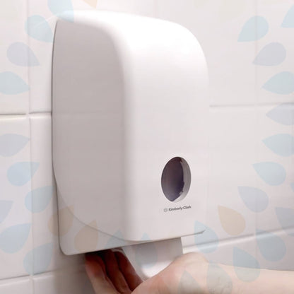 A Kleenex® Gentle Wash Lotion cassette from Kimberly-Clark GmbH (transparent, 1 L) is depicted on a white tiled wall, with a hand pulling a towel from the bottom and a small round window on the front. Available in packs of 6 cassettes.