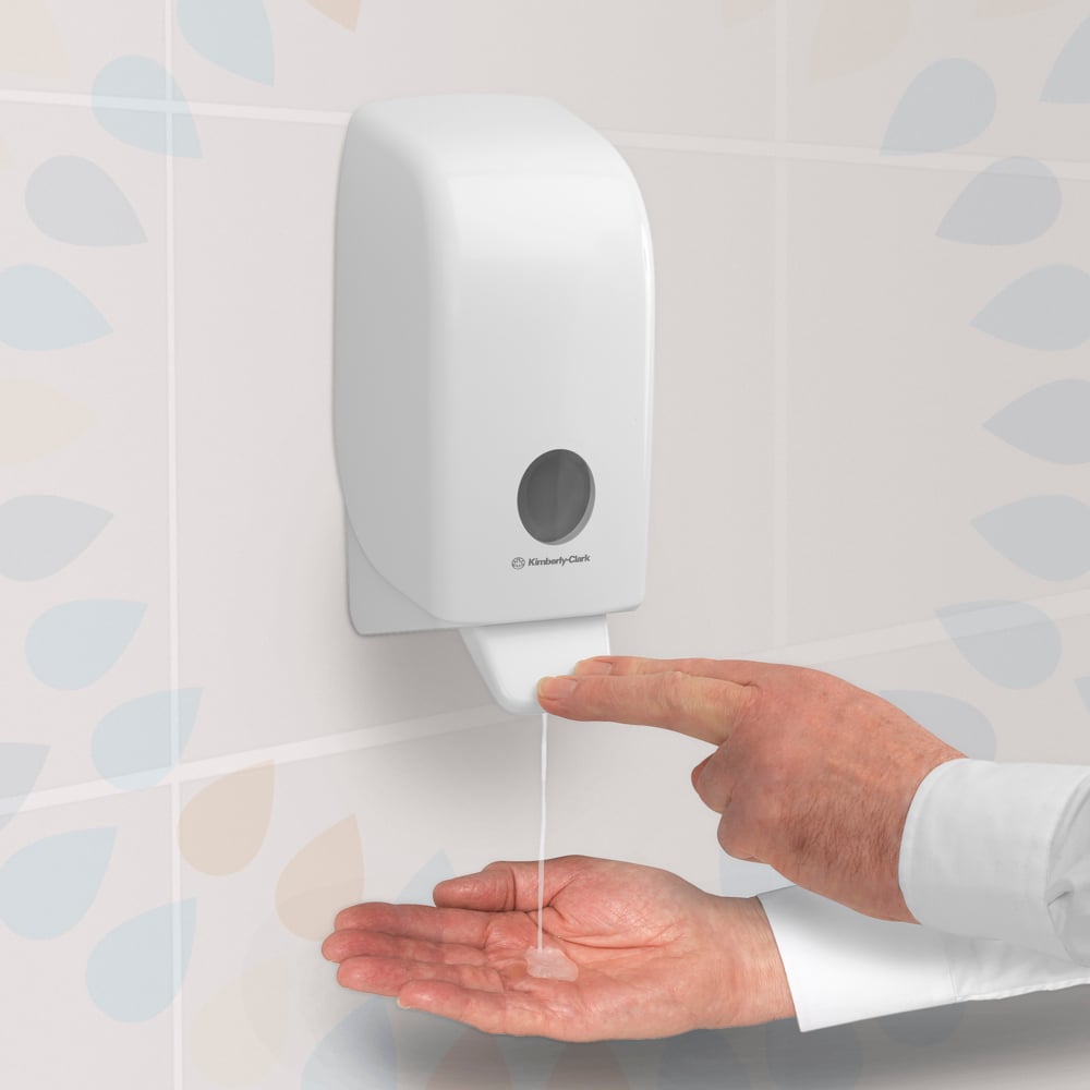 Eine Person drückt auf einen transparenten Kleenex® Extra Gel Händedesinfizieren-Spender der Kimberly-Clark GmbH, der an einer gefliesten Wand angebracht ist, und gibt das Handdesinfektionsgel aus einer 1-Liter-Kassette auf ihre offene Handfläche ab.