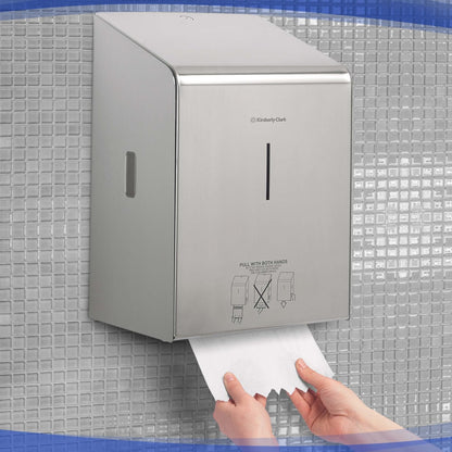 A person pulls paper towels from a Kimberly-Clark Professional™ stainless steel roll towel dispenser by Kimberly-Clark GmbH, which is mounted amidst small gray square tiles on the wall.