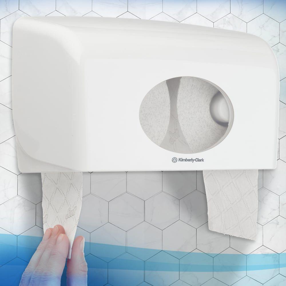 A person pulls FSC-certified Scott® Essential™ Toilet Paper 8517 2-ply from Kimberly-Clark GmbH from a white wall dispenser in front of a hexagon-tiled bathroom wall.