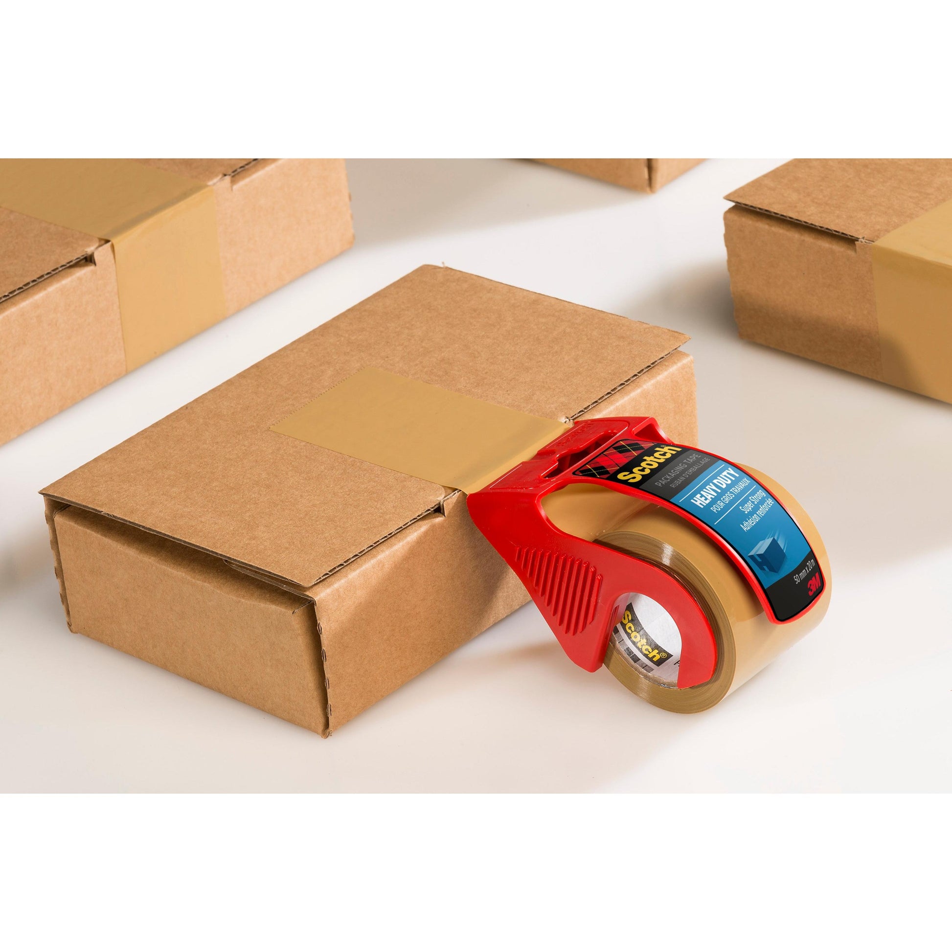 Eine Rolle Scotch® Extra starkes Verpackungsklebeband, Braun, 48 mm x 20,3 m von 3M Deutschland GmbH wird in einem roten Handabroller zum Verschließen eines braunen Kartons genutzt. Weitere versiegelte Boxen stehen im Hintergrund auf weißer Fläche.