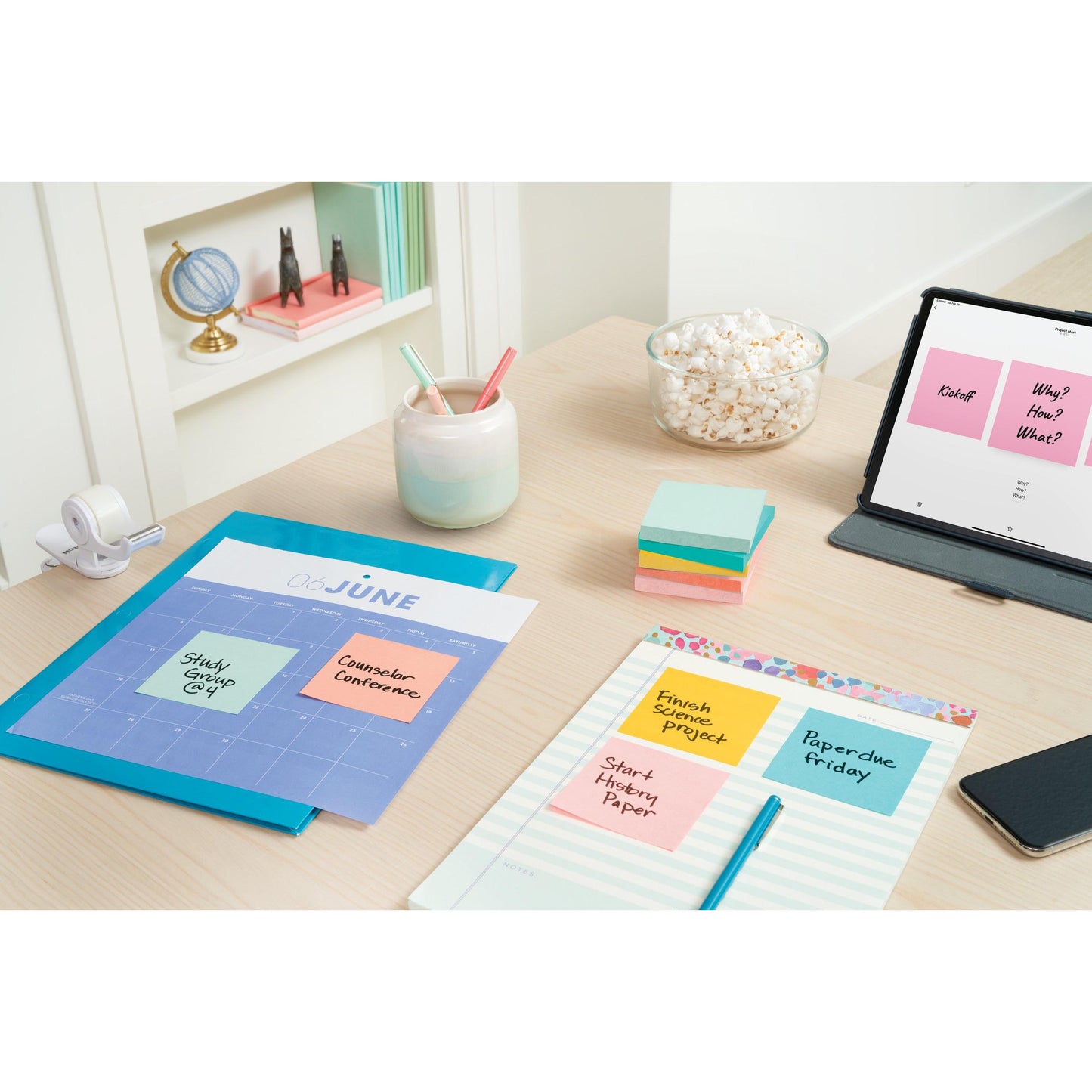 Ein aufgeräumter Schreibtisch zeigt einen Kalender, einen Planer, Ordner, Stifte in einem Halter, eine Popcorn-Schale, einen Klebebandabroller, ein Tablett mit Notizen und Post-it® Notes (76 mm x 76 mm, 100 Blatt/Block) von 3M Deutschland GmbH; in Regalen stehen Bücher und im Hintergrund ein Globus.