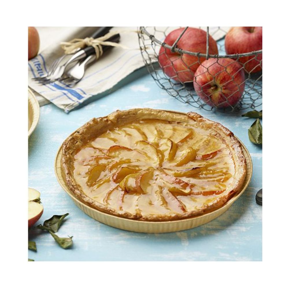 A baked apple pie with spiral apple slices lies on a blue surface, resting on PAPSTAR 25 baking molds "pure" round brown from PAPSTAR GmbH. In the background, there is a basket of apples and a striped towel with cutlery.
