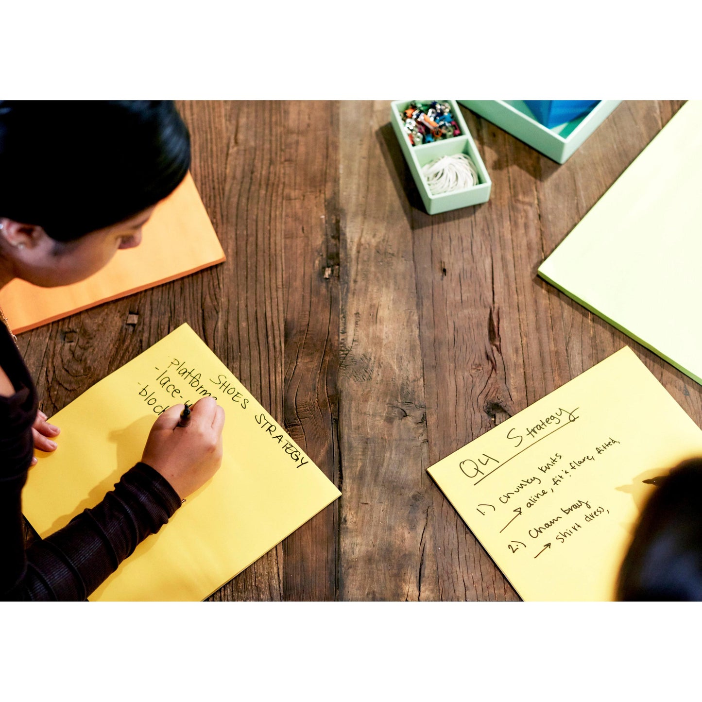 Zwei Personen sitzen an einem Holztisch und schreiben auf 3M Post-it® Super Sticky Big Notes BN11-EU, Gelb (279 x 279 mm, 30 Blatt/Block, 100% PEFC) und brainstormen Ideen wie "PHOTOSHOOT SHOES STRATEGY" und "Q4 Strategy". Gelbe Haftnotizen und Marker liegen bereit.