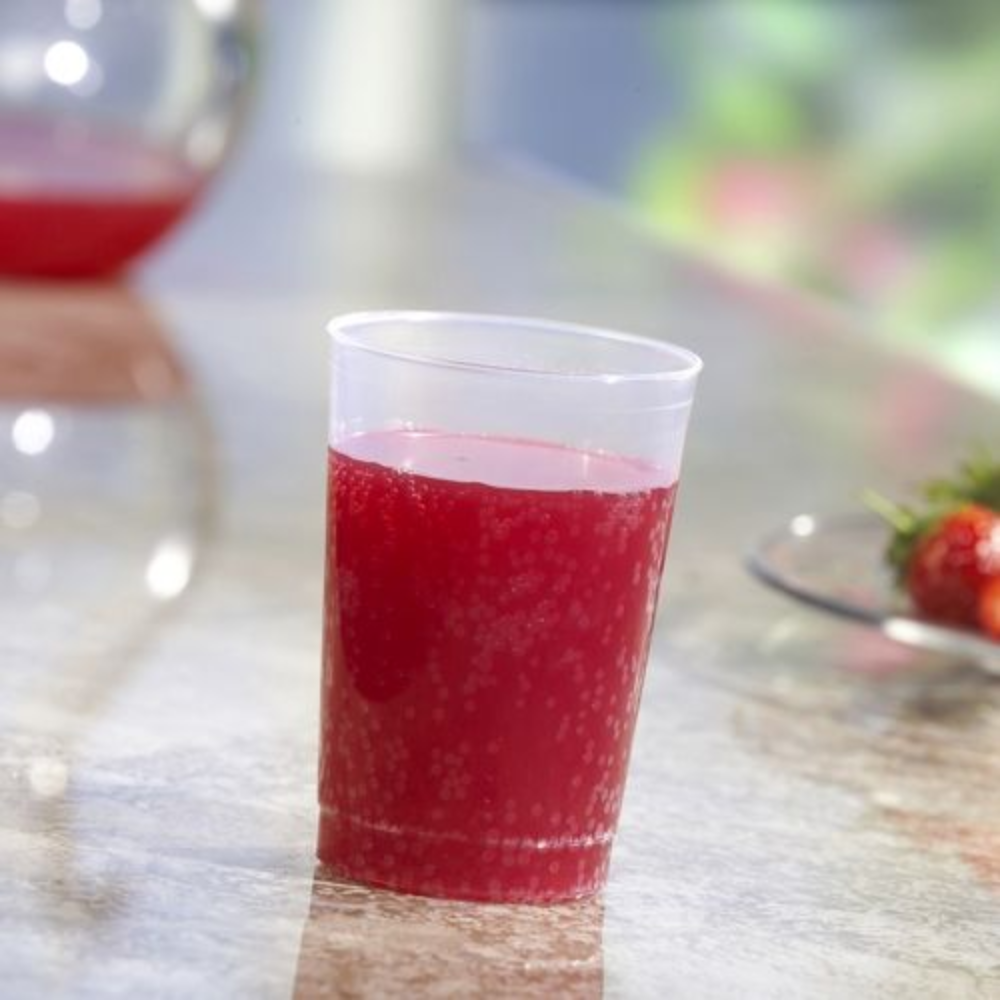 A Starpak 40 reusable drinking cup made of translucent, unbreakable polypropylene from Starpak, filled with a red drink, stands on a shiny surface. In the background, another cup and a plate of strawberries can be seen blurred.