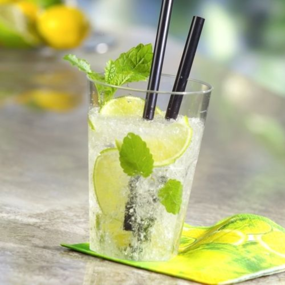 A Starpak reusable plastic cup filled with a lime-mint cocktail, garnished with fresh lime slices and mint leaves. Inside are two black straws, placed on a napkin with a lemon pattern. The blurred background shows more limes and green plants.