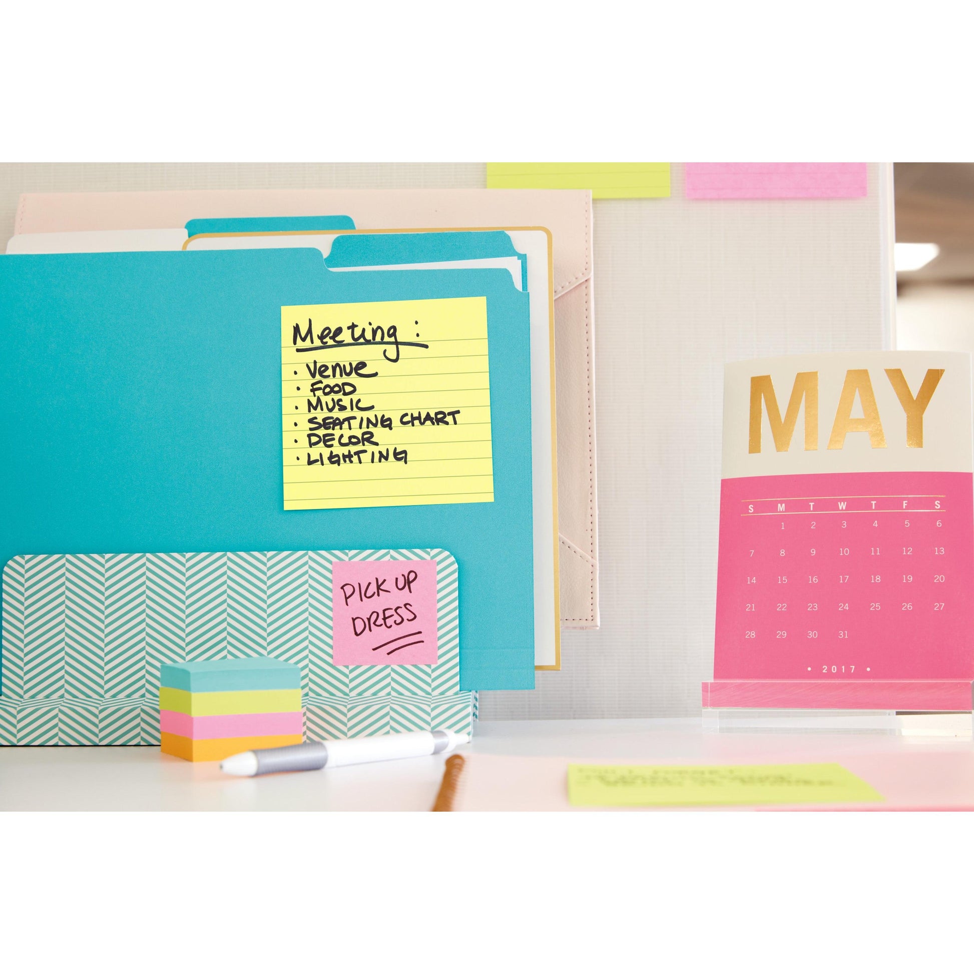 Ein Schreibtisch mit bunten Ordnern, einem Stapel Haftnotizen und einem rosa-goldenen Kalender für Mai 2017. Auf den Post-it® Super Sticky Notes (101 mm x 101 mm, 90 Blatt, PEFC) der 3M Deutschland GmbH sind Besprechungsthemen und "PICK UP DRESS.
