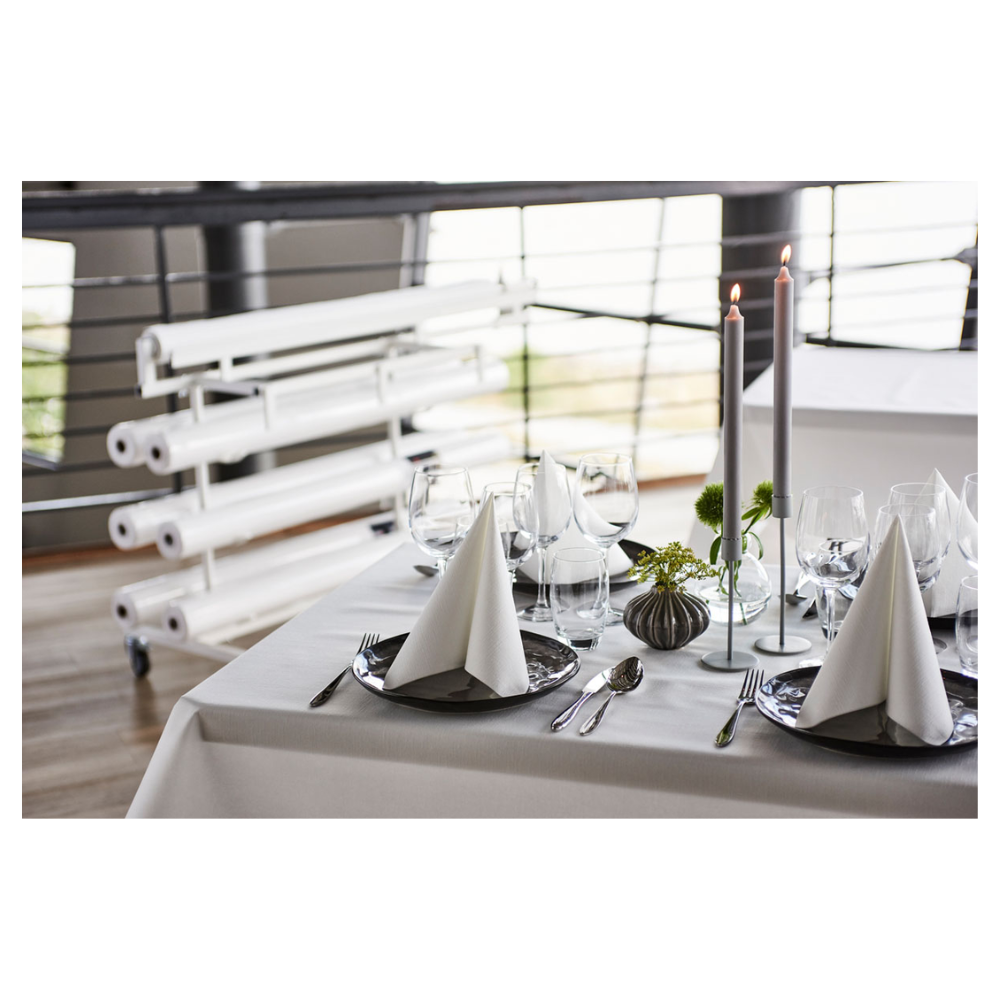 On a beautifully set dining table stands a fresh, white Duni Dunicel® tablecloth roll from Duni GmbH, offering a fabric-like texture. Triangular folded napkins on dark plates accompany wine glasses. Tall candles and small floral arrangements serve as table decorations, with rolled canvases visible in the background.