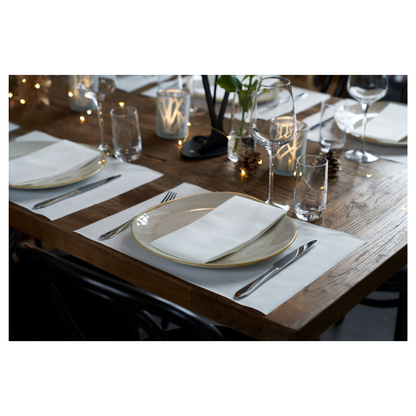 A set dining table made of wood with white placemats, plates, glasses, and cutlery. Folded Duni Dunilin® napkins 48 x 48 cm white from Duni GmbH add charm. Burning candles and small decorative lights enhance the cozy atmosphere.
