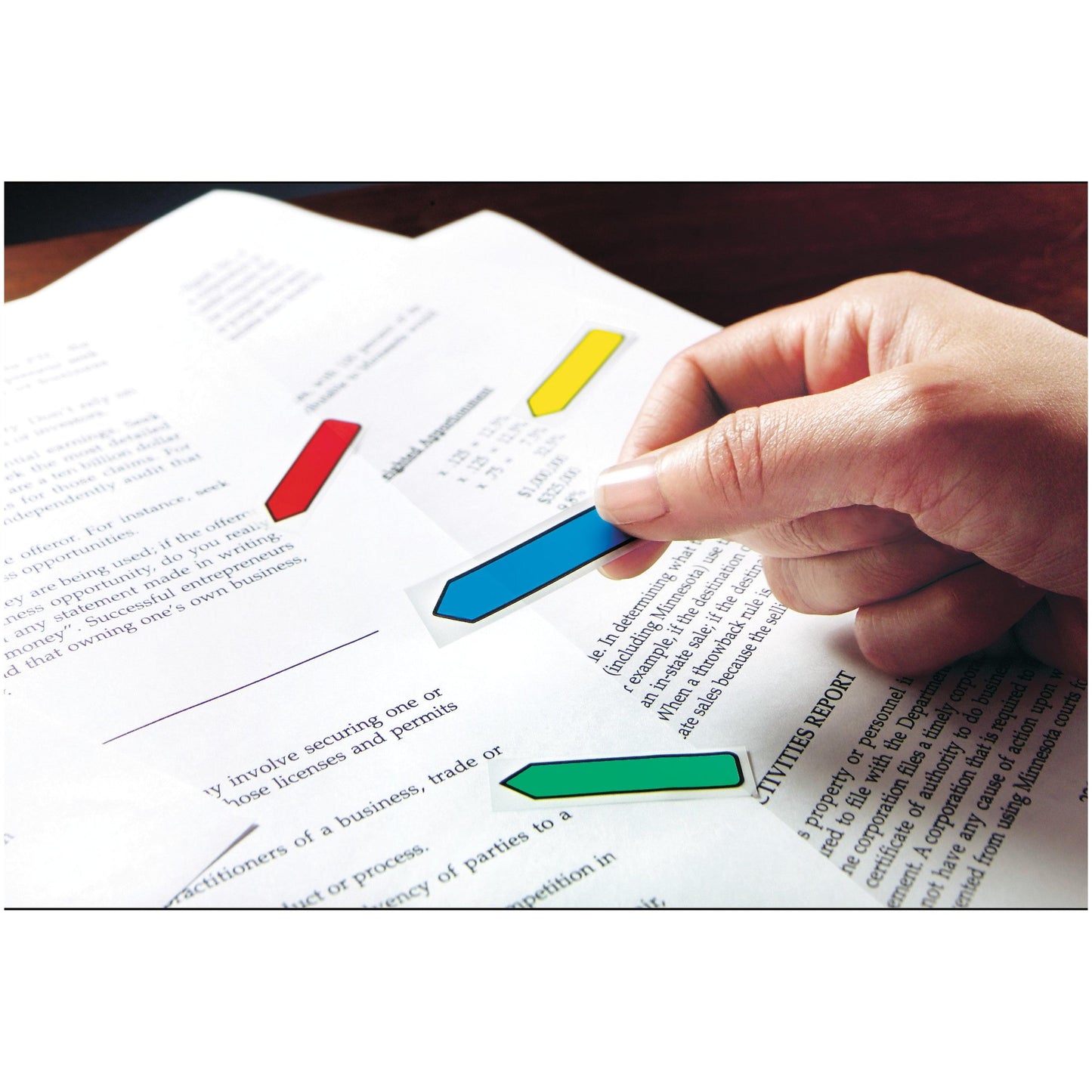 A hand sticks a green Post-it® Index Arrow (11.9 mm x 43.2 mm) from 3M Deutschland GmbH onto a document that is already marked with blue, red, and yellow tabs; the text of the document is partially visible.
