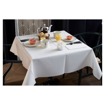 The breakfast table is elegantly set with a white tablecloth, two chairs, and a vase with a geometric pattern. On the table are grapefruit halves, pastries, tea, and orange juice. A Duni Dunilin® napkin (48x48 cm) from Duni GmbH adds charm and functionality to the atmospheric morning setting.
