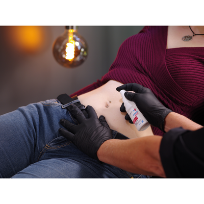 A person is wearing black gloves and cleaning another person's navel with Schülke octenisept® wound disinfection spray 50 ml from Schülke & Mayr GmbH. The recipient is wearing a burgundy top and blue jeans. In the background, a bright light bulb can be seen.