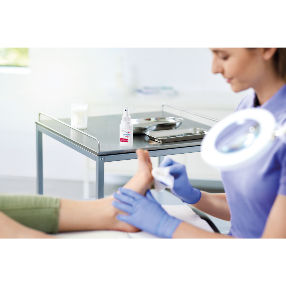 A person with blue gloves is cleaning another person's foot in a clinic. The focus is on infection protection. On the table are a 50 ml bottle of Schülke octenisept® wound disinfection spray from Schülke & Mayr GmbH and a metal tray with tools.
