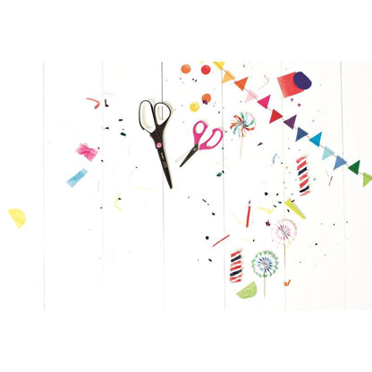 A Scotch™ titanium scissors (20 cm) from 3M Deutschland GmbH, colorful paper scraps, confetti, lollipops, candles, and a small rainbow banner are scattered on a white wooden surface, creating a festive, creative atmosphere.