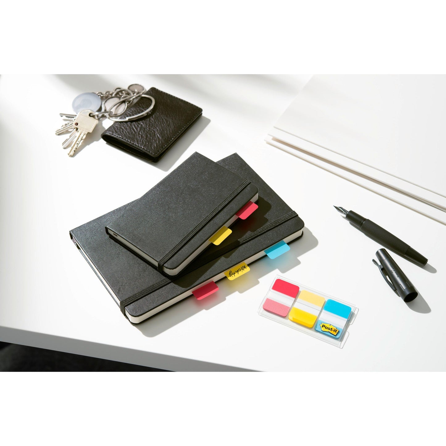 A set of black notebooks with Post-it® Index Strong Filing Tabs from 3M Deutschland GmbH in various colors, a wallet, keys, white paper, a black pen, and a marker lie on a white desk.