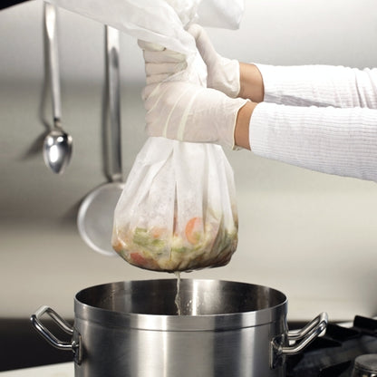 A person with white gloves uses Starpak 10 straining and filter cloths 100 cm x 100 cm white to efficiently drain vegetables into a stainless steel pot on the stove, while kitchen utensils hang on the wall in the background.