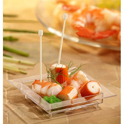 A small square transparent dish contains imitation crab, green garnish, and two PAPSTAR 100 Fingerfood - Picks from PAPSTAR GmbH. Blurred vegetables and other seafood appear in the background.