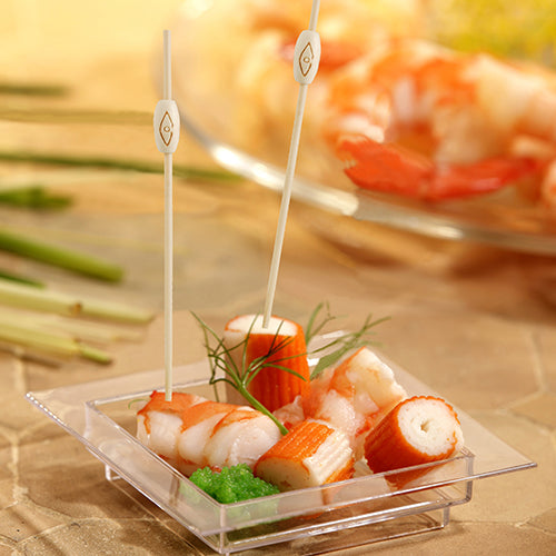 A square, transparent dish contains imitation crab, shrimp, and green roe with herbs. Two PAPSTAR 100 Fingerfood - Picks from PAPSTAR GmbH are inserted into the seafood. Blurred shrimp appear in the background.