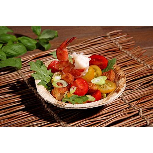 A small bowl with cooked shrimp, cherry tomatoes, croutons, arugula, and spring onions is served on a woven base in the PAPSTAR 25 bowls, palm leaf "pure" from PAPSTAR GmbH - an eco-friendly choice for sustainable events. Basil leaves add a fresh touch.