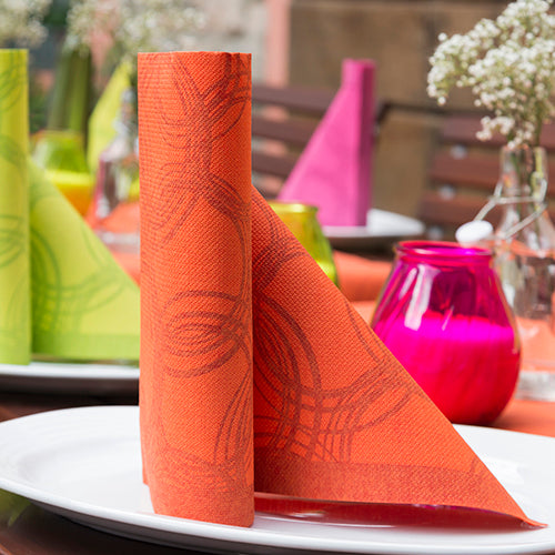A neatly folded orange PAPSTAR 50 napkins "ROYAL Collection" 1/4-fold 40 x 40 cm nectarine napkin stands upright on a white plate, with colorful ROYAL Collection decor and blurred flowers in the background.