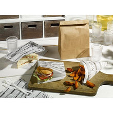 A sandwich and potato wedges in a PAPSTAR cone bag "Newsprint", greaseproof, 32.5x23 cm (parchment substitute), are served on a wooden board with cups, plastic forks, and napkins with newspaper motifs. Brand: PAPSTAR GmbH.
