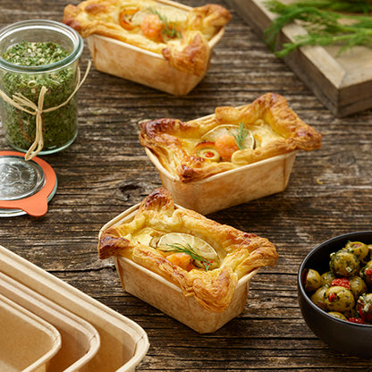 Three savory pies with golden puff pastry lie on a table next to a glass of green seasoning, marinated olives, herbs, and a stack of PAPSTAR baking molds "pure" rectangular (4x6.5x11 cm, brown) from PAPSTAR GmbH (50 pieces) in shrink wrap.