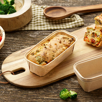 A vegetable loaf lies in a PAPSTAR 25 baking molds "pure" rectangular brown by PAPSTAR GmbH on a wooden board, with another empty rectangular mold next to it. Broccoli pieces and a checkered tablecloth are also on the table.