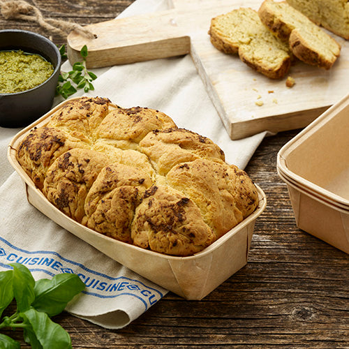 A loaf of hearty bread in a PAPSTAR 25 baking molds "pure" rectangular brown by PAPSTAR GmbH lies on a cloth next to fresh herbs, while in the background, sliced bread and a jar of green pesto are presented on a wooden board.