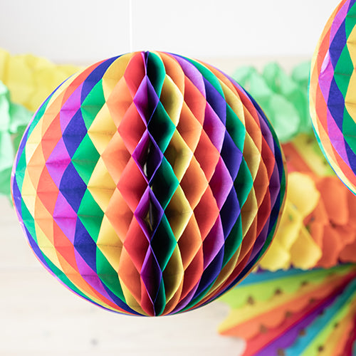 A close-up of the flame-retardant honeycomb ball from PAPSTAR GmbH shows its bright rainbow colors, with another honeycomb ball and green-yellow paper garlands blurred in the background.