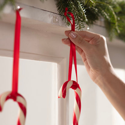Eine Hand hängt eine rot-weiße Zuckerstange an einem roten Band auf einer weißen Fläche auf, mit Command™ Mini-Haken, transparent, mit transparenten Strips, 17006CLRFGN von 3M Deutschland GmbH. Ein weiterer Zuckerstab hängt bereits daneben an grünem Tannengirlande.
