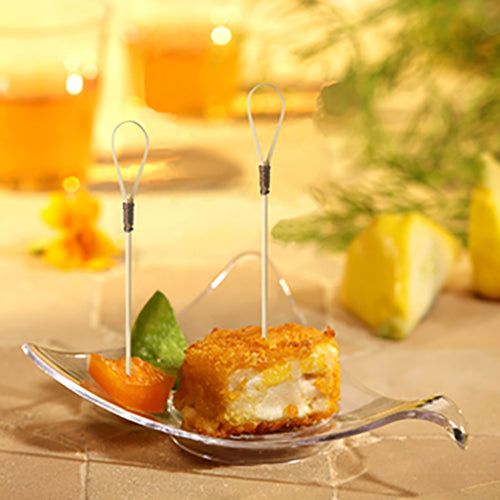 A glass bowl presents a breaded, fried appetizer cube with a PAPSTAR finger food picker "Loop" (10 cm), an orange segment, and a green garnish. In the background, lemon wedges and two drinks from PAPSTAR GmbH blur.