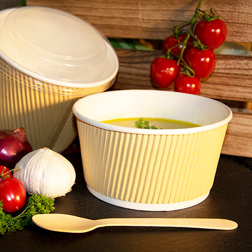 A yellow Starpak soup cup with soup and herbal garnish stands next to a wooden spoon, garlic, red onions, cherry tomatoes, and parsley; in the background, a round Starpak 25 lid for R-PET soup cups can be seen.