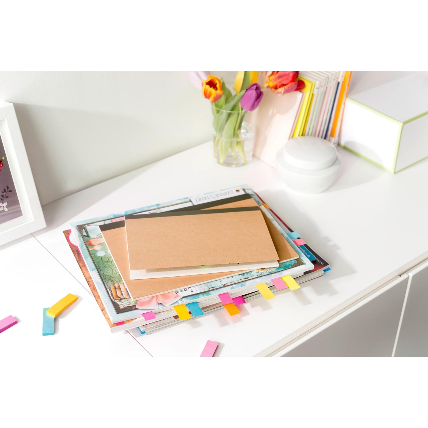 Auf einem weißen Schreibtisch liegen Zeitschriften und ein Notizbuch mit den Post-it® Page Markern (Verschiedene Farben, 15 mm x 50 mm, Promo Pack: 6 Blöcke + 3 Gratis) der 3M Deutschland GmbH, dazu eine Blumenvase, aufrecht stehende Bücher und verstreute farbige Seiten markieren Tabs.
