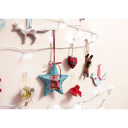 An einer beigen Wand hängen weiße Lichterketten und festliche Ornamente - darunter ein silberner Fuchs, ein blauer Stern, ein kariertes Herz und eine weiße Taube - an transparenten Command™ Dekorations-Clips der 3M Deutschland GmbH.