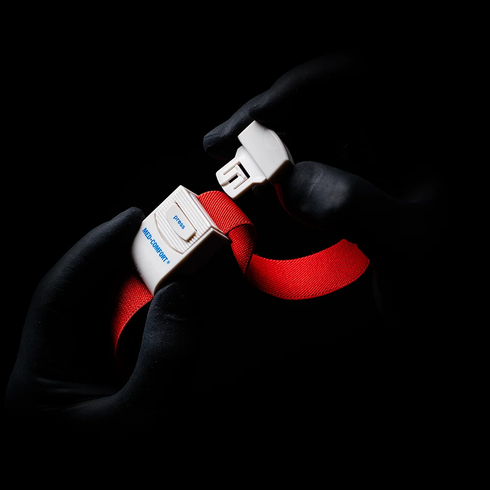 Behandschuhte Hände, die an einen AMPri MED-COMFORT Venenstauer erinnern, greifen vor einem schattigen Hintergrund die Schnalle eines leuchtend roten Sicherheitsgurts. Auf der Schnalle sind das Wort „Blue“ und der Buchstabe „B“ zu sehen.